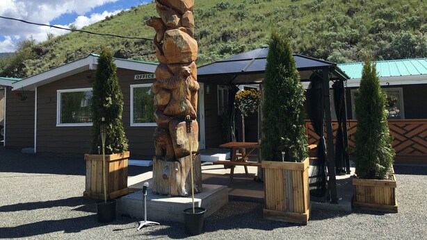 A rustic wooden sculpture in front of a building labeled "OFFICE" with a patio area, potted plants, and a hill in the background. - Sunset Motel (Cache Creek)