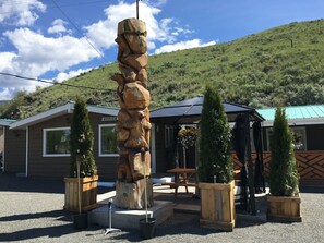 Une sculpture rustique en bois se trouve devant un bâtiment portant l’inscription « BUREAU », avec un espace patio, des plantes en pot et une colline en arrière-plan.