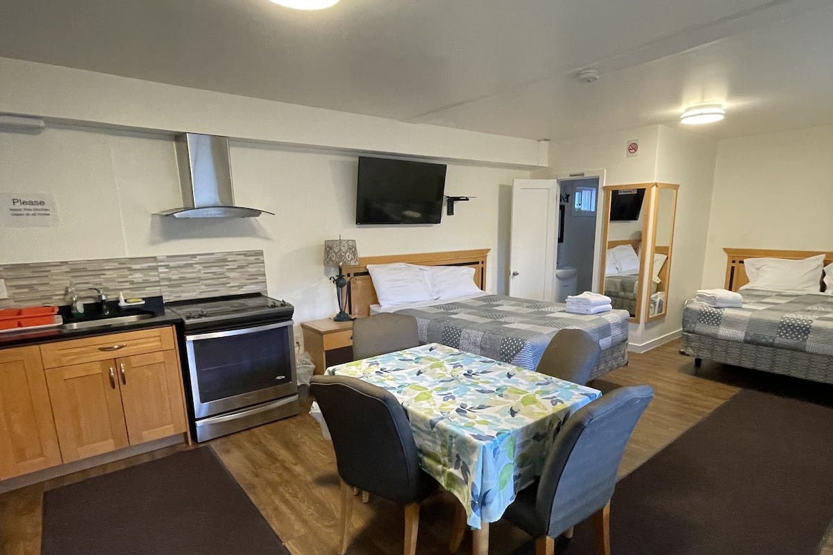 Habitación de hotel compacta con kitchenette, un comedor con mesa y sillas, una cama con cabecera y un televisor fijado a la pared.