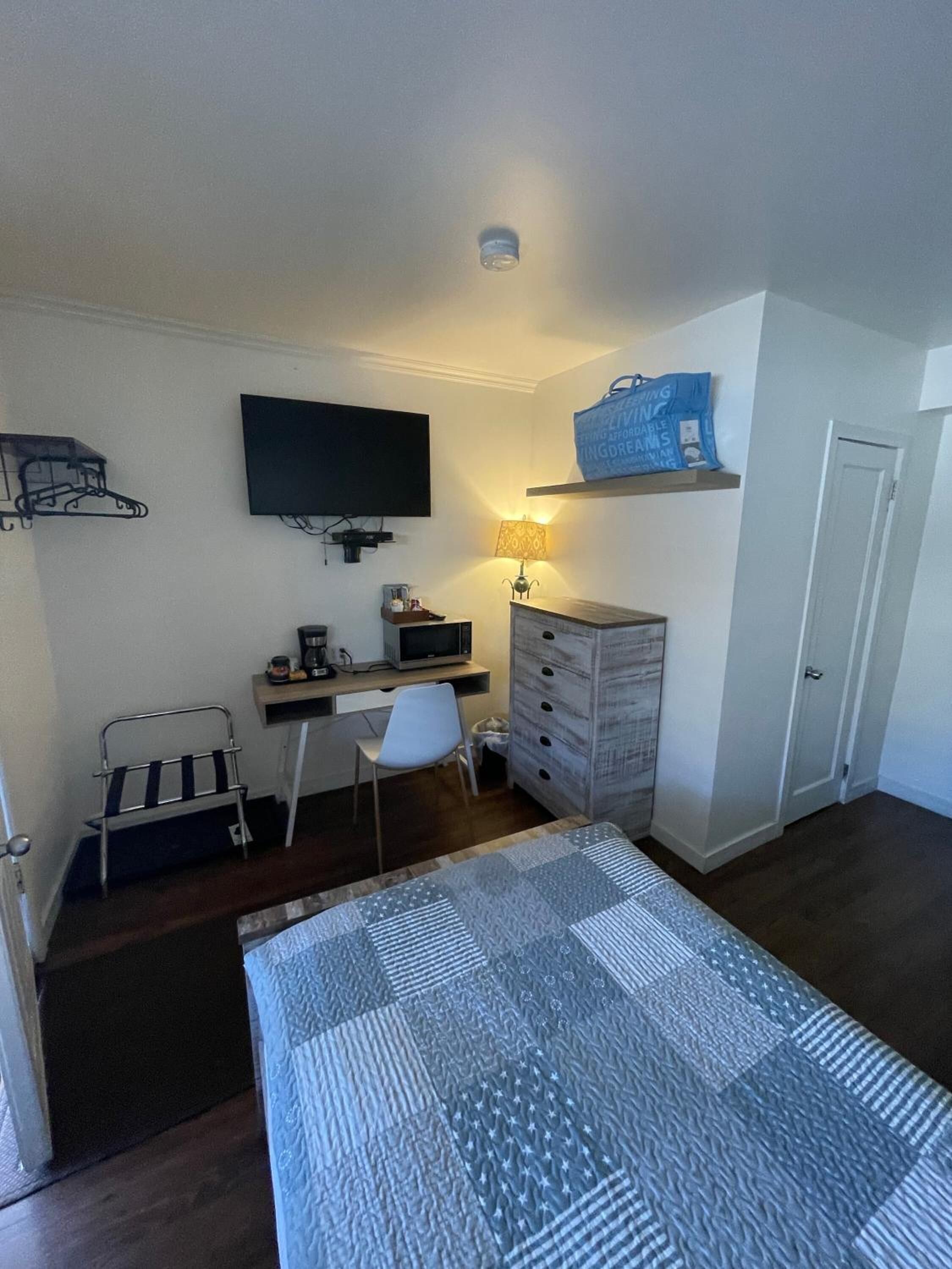 Habitación de hotel pequeña con cama, escritorio con ordenador, televisión, silla, armario y un estante con un bolso.