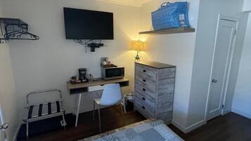 Habitación de hotel pequeña con cama, escritorio con ordenador, televisión, silla, armario y un estante con un bolso.