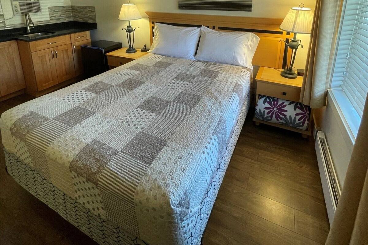 Habitación de hotel con cama, mesitas de noche, lámpara, ventana con persianas, zona de cocina y un cuadro en la pared.