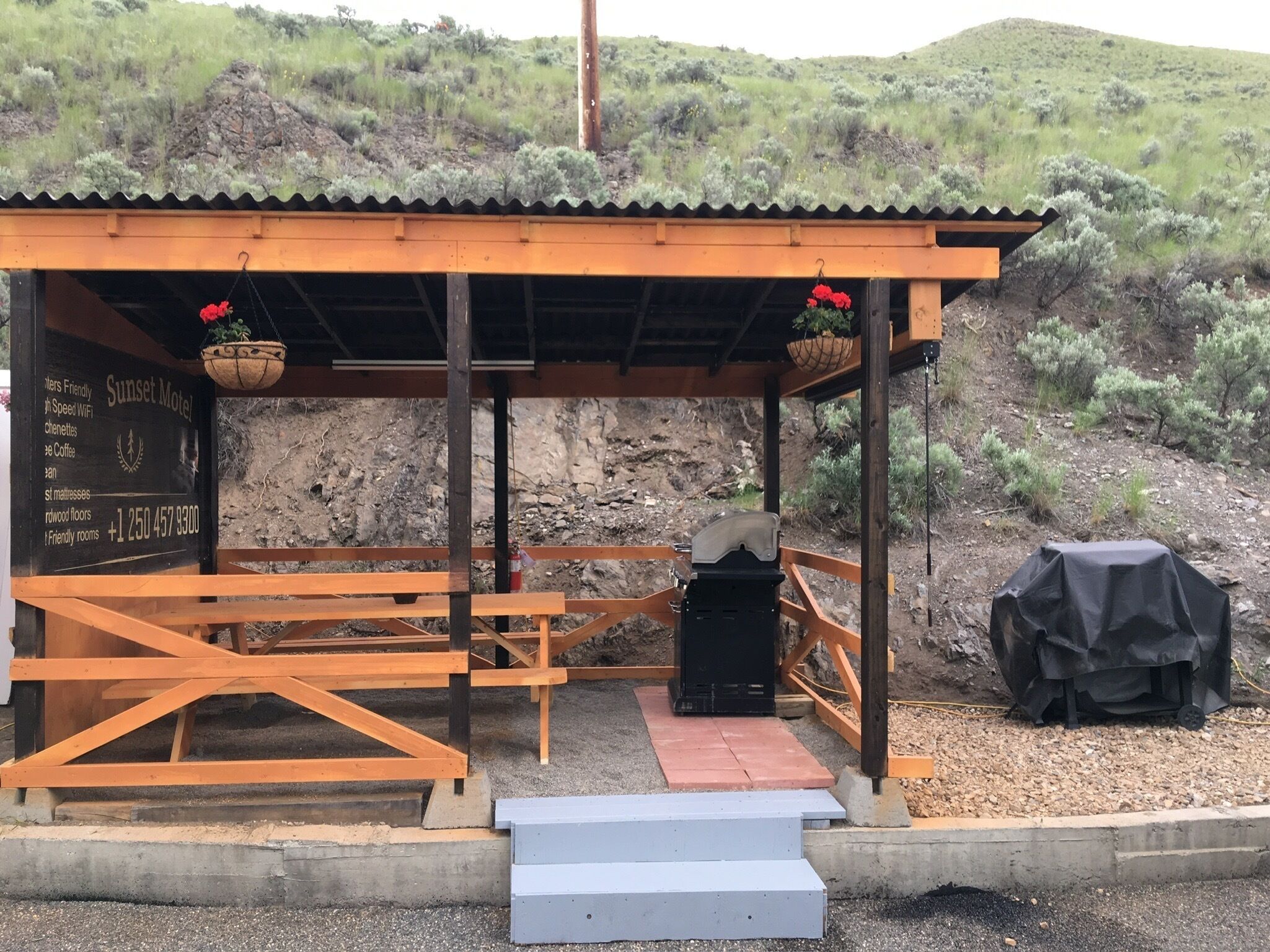 A rustic outdoor seating area with wooden benches and a covered shelter, featuring a chalkboard sign with amenities and contact information.