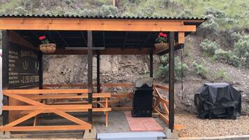 A rustic outdoor seating area with wooden benches and a covered shelter, featuring a chalkboard sign with amenities and contact information.