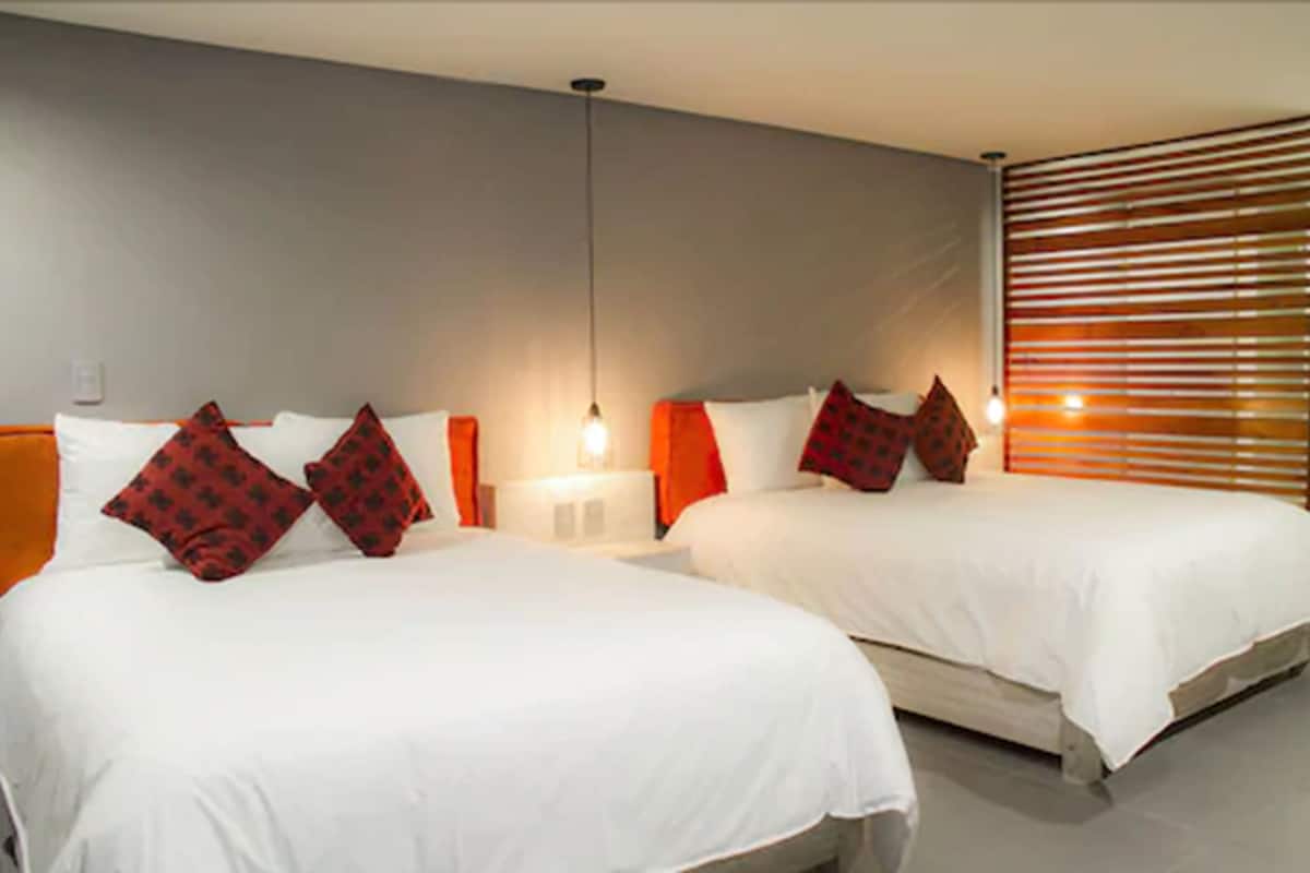 A hotel room with two beds, each with white bedding and red patterned pillows. There are bedside lamps and a window with blinds.