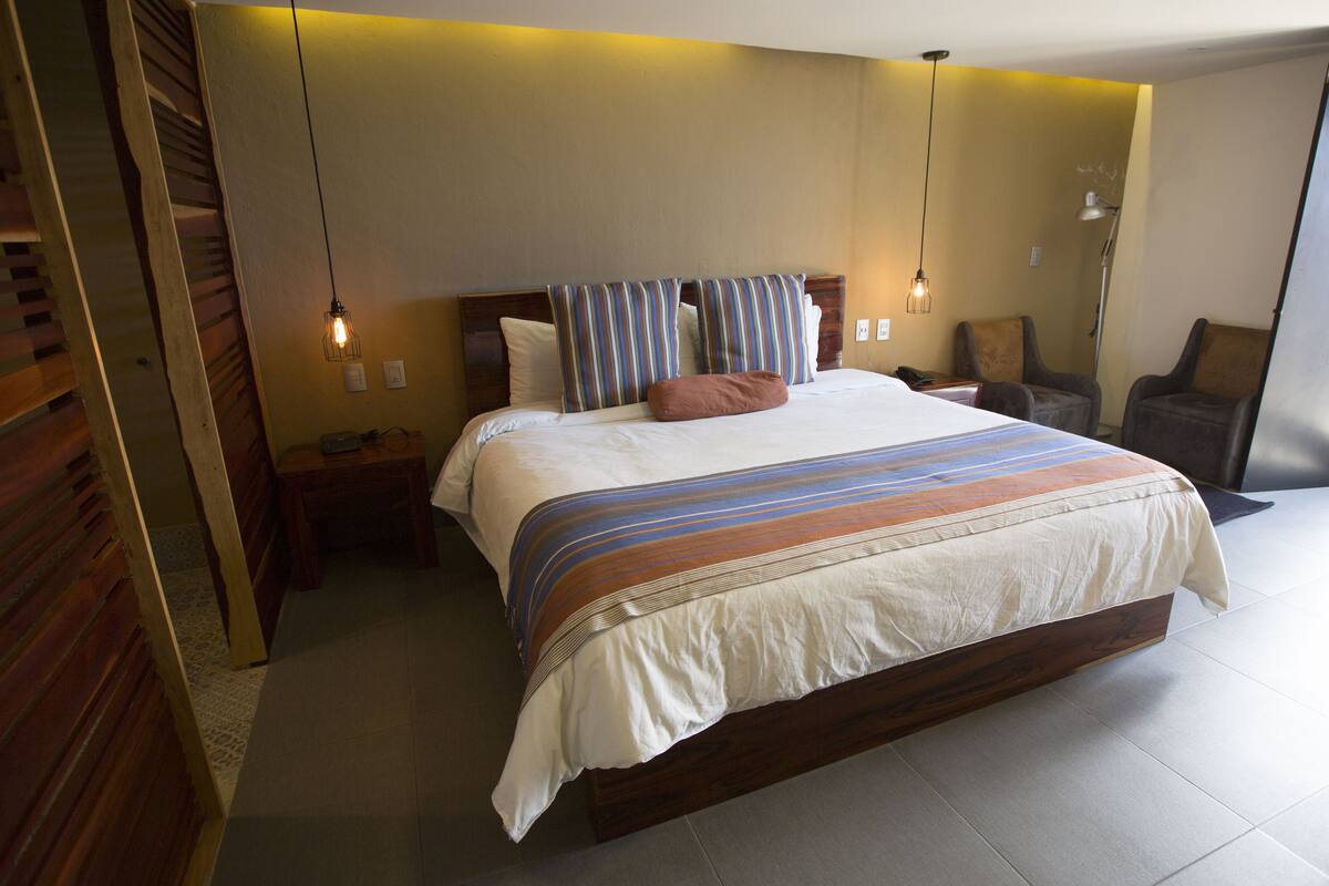 A modern hotel room with a large bed, wooden headboard, and a striped throw blanket. There are two bedside tables with lamps, a chair, and a large window.