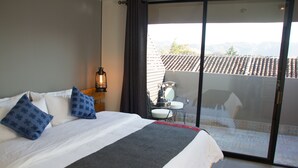 A bedroom with a large bed, a bedside table with a lantern, a chair, and a view of a tiled roof and mountains through a sliding door.