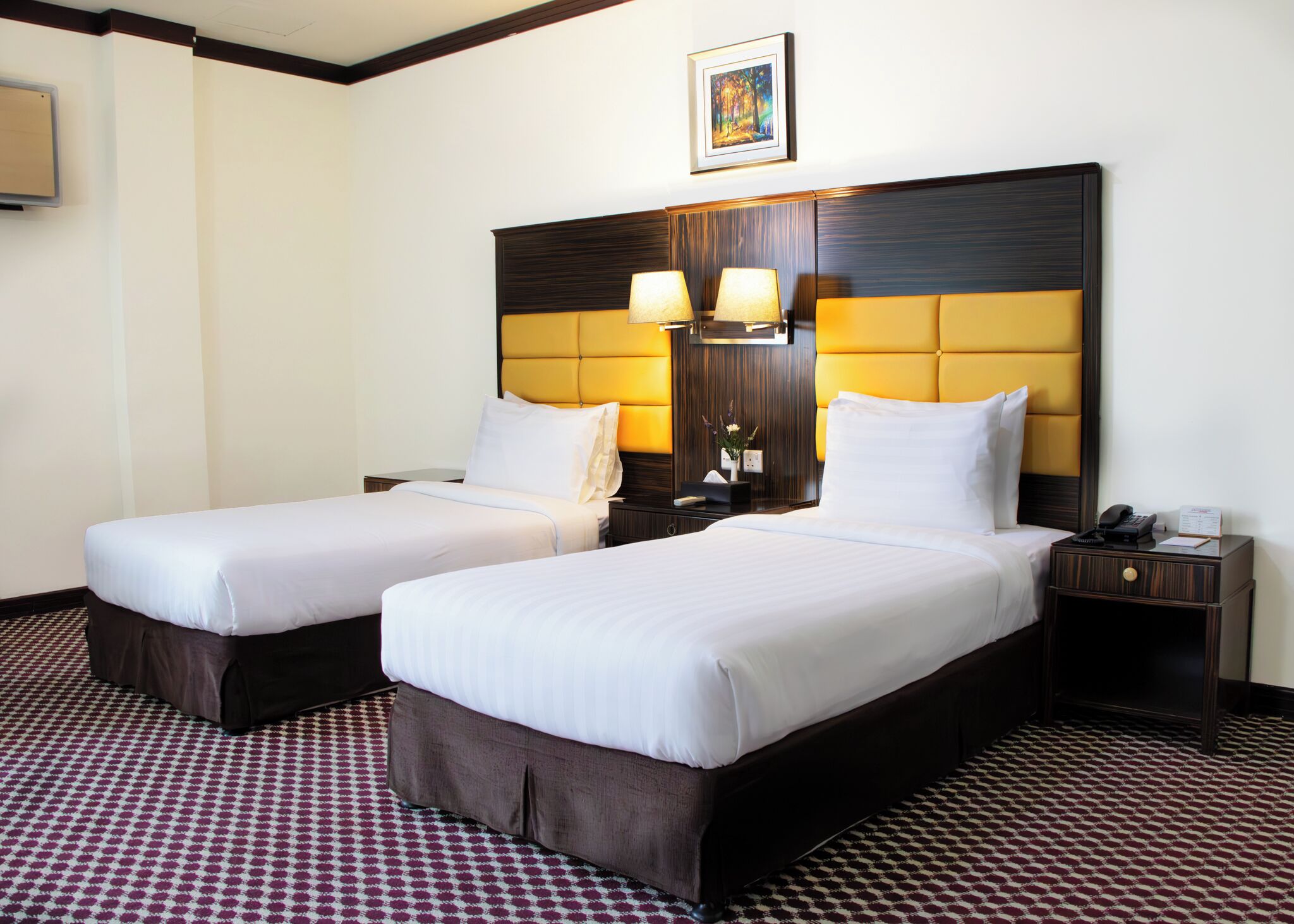 A hotel room with two beds, a large wooden headboard, bedside lamps, a small table with a phone, and a patterned carpet.
