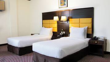 A hotel room with two beds, a large wooden headboard, bedside lamps, a small table with a phone, and a patterned carpet.