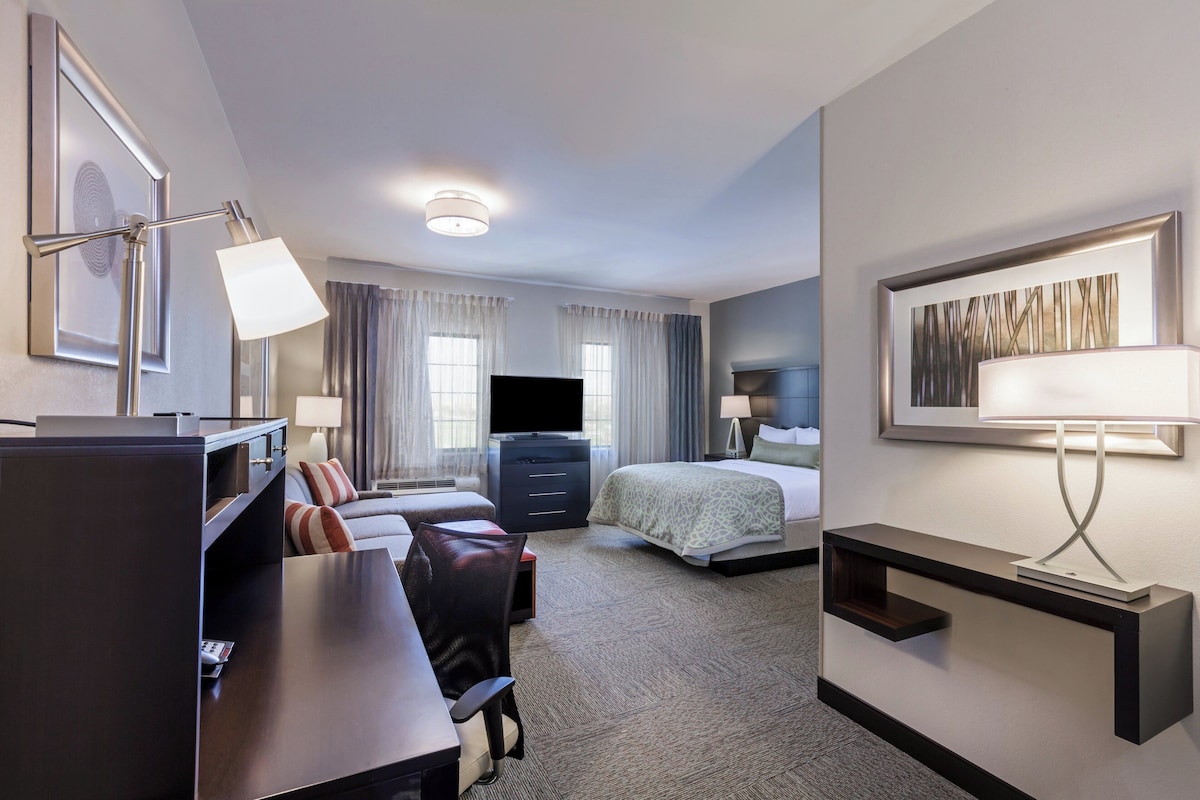 Una habitación de hotel con cama, escritorio, silla, televisor y obras de arte en las paredes.