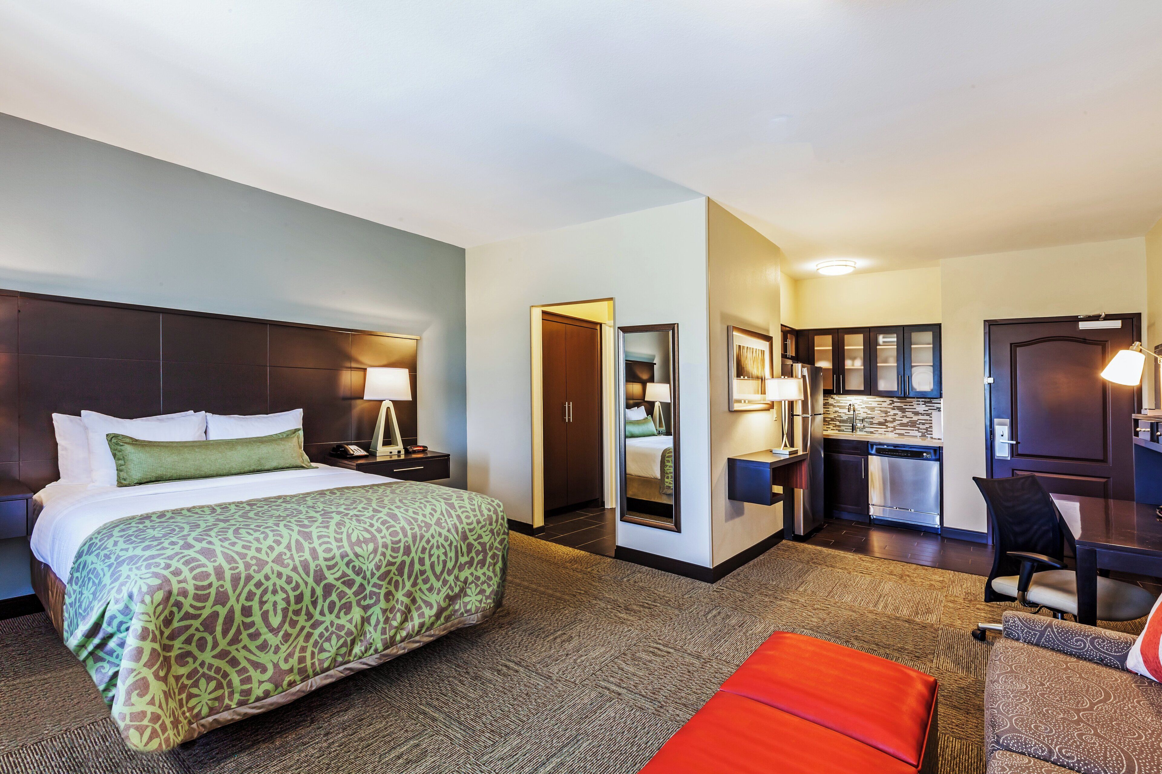 A hotel room with a bed, a desk, a chair, a kitchenette, and a bathroom.