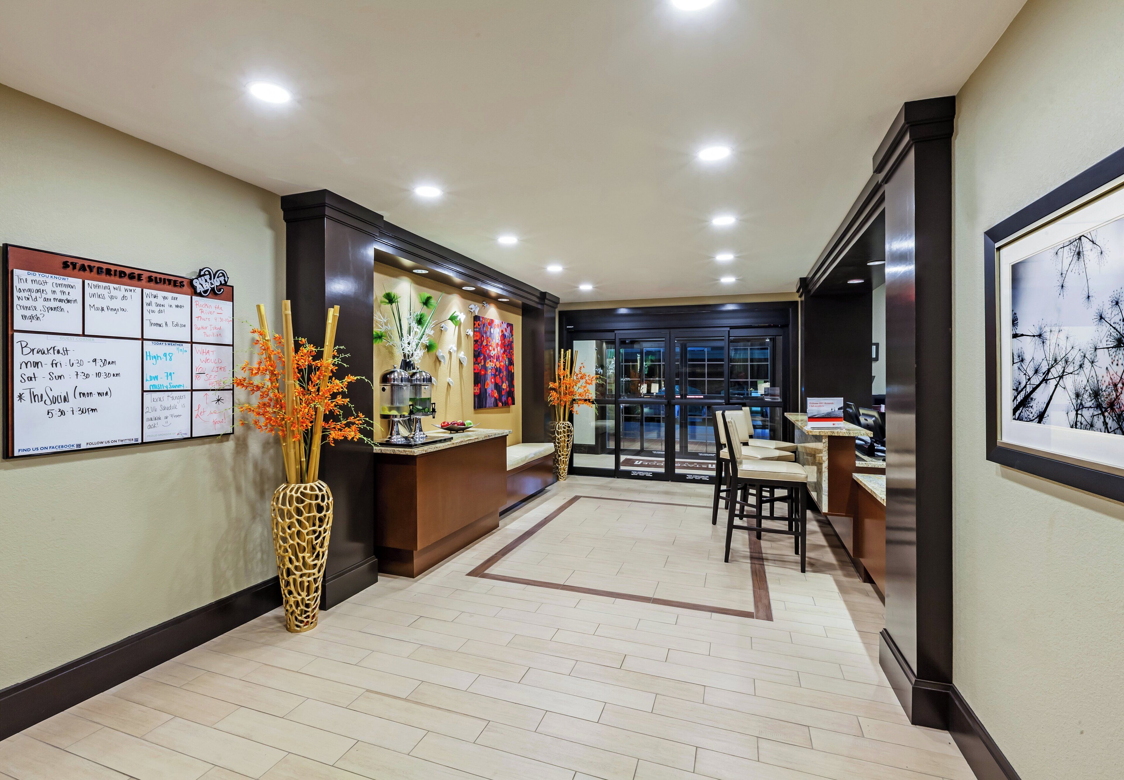 A hotel reception area with a reception desk, a bulletin board with information, a vase with flowers, and a framed picture on the wall.