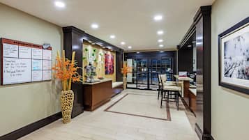 A hotel reception area with a reception desk, a bulletin board with information, a vase with flowers, and a framed picture on the wall.