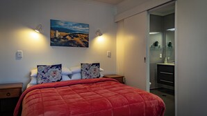 A bedroom with a large bed, two nightstands, a painting on the wall, and a bathroom visible through an open door. - Amber Court Motel (Te Anau)