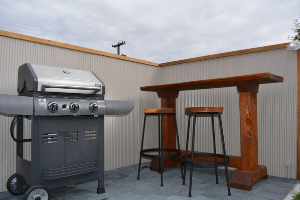 A barbecue grill, two stools, and a wooden table against a corrugated metal wall.
