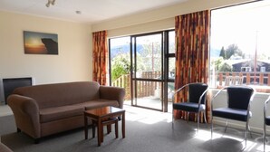 A living room with a sofa, a small table, and two chairs. There is a TV on the left and a large window with curtains. - Amber Court Motel (Te Anau)