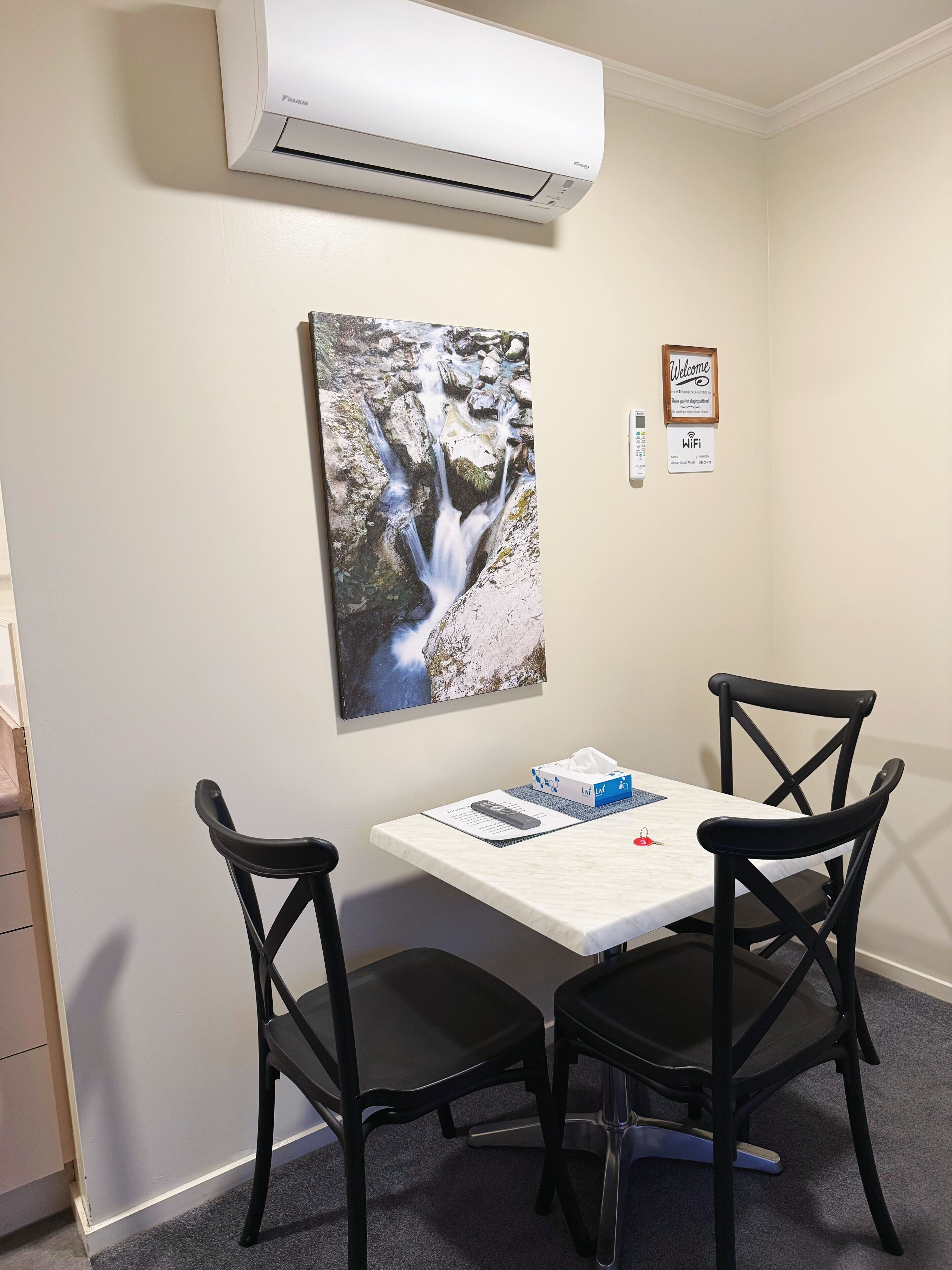 A room with a white air conditioning unit, a painting of a waterfall, a table with a tissue box, and two black chairs.