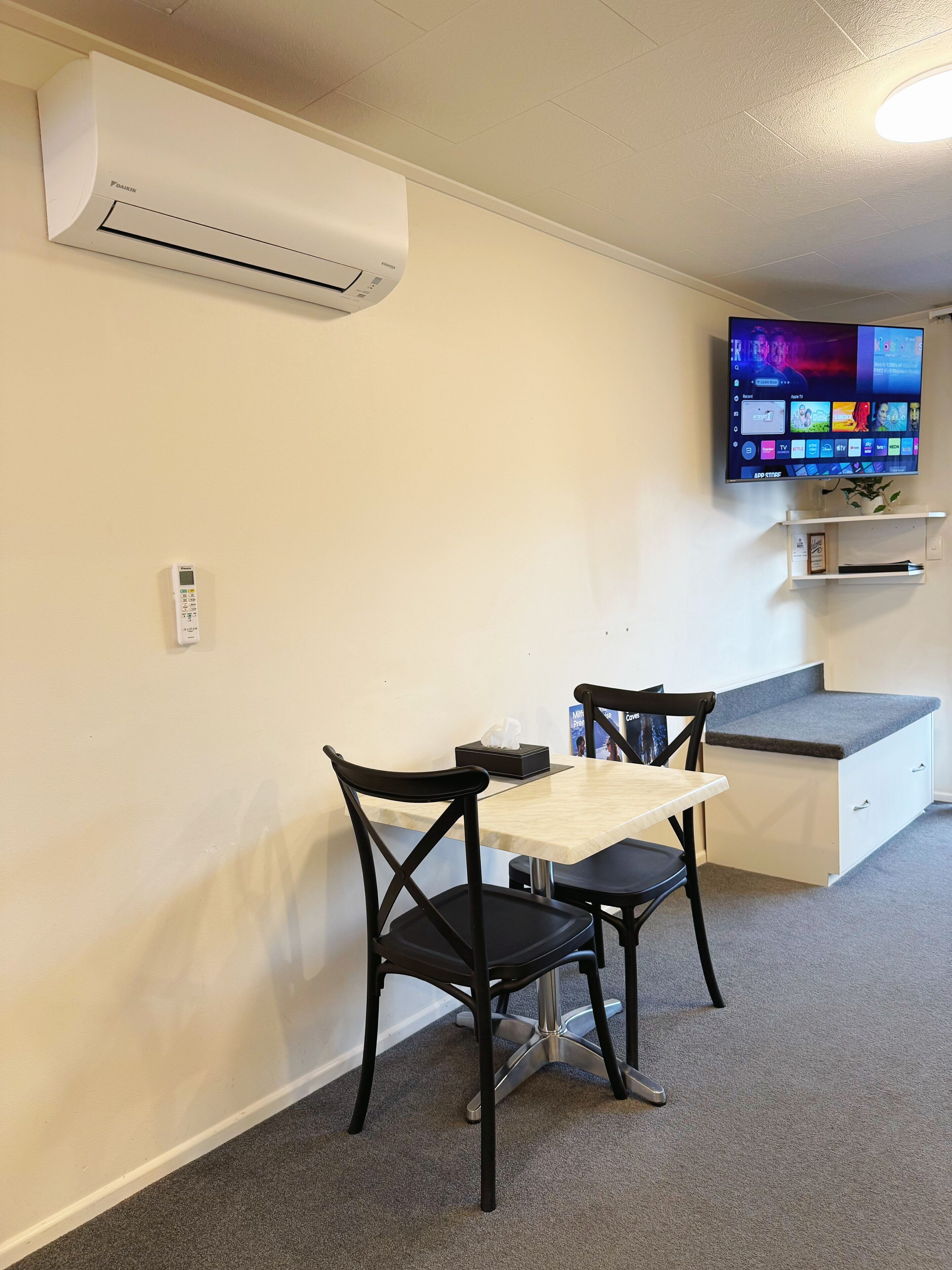 A room with a table and chairs, an air conditioning unit, a television, and a small shelf with items.