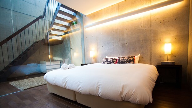 A modern bedroom with a large bed, a wooden staircase, and a concrete accent wall. - Full Circle (Kutchan)