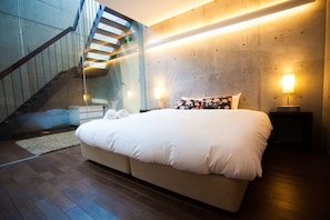 A modern bedroom with a large bed, a wooden staircase, and a concrete accent wall. - Full Circle (Kutchan)
