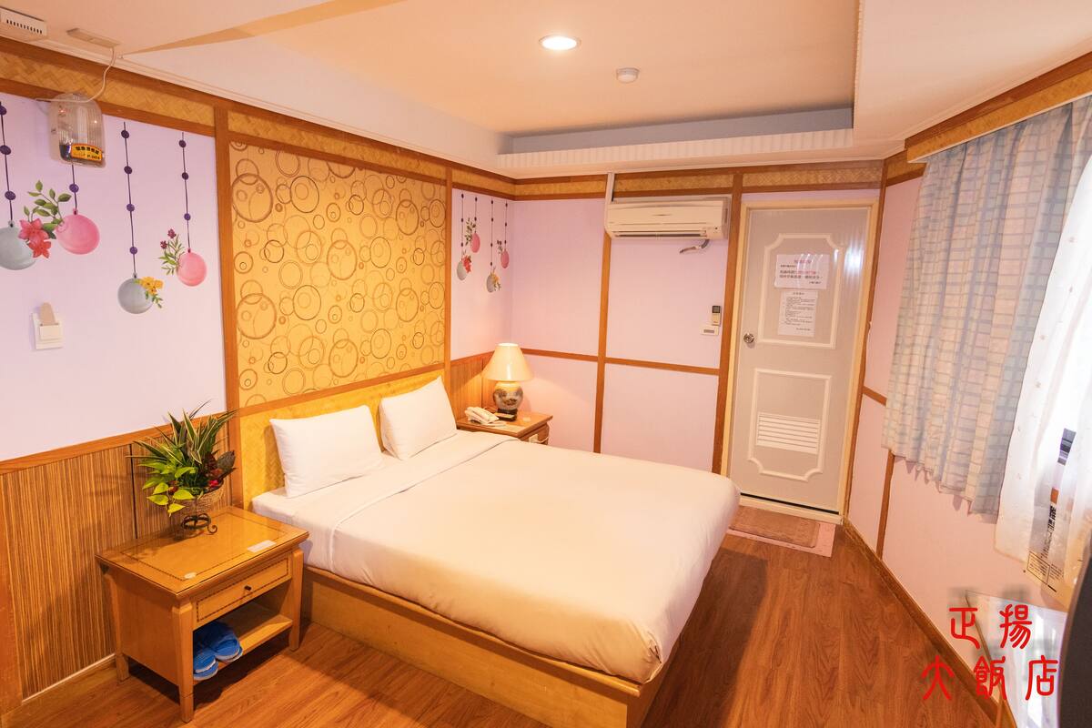 A hotel room with a wooden bed, a nightstand with a lamp, a wall-mounted air conditioner, and a door with a sign.