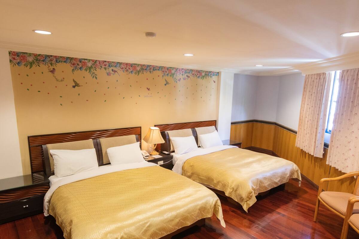A hotel room with two beds, wooden headboards, a nightstand with a lamp, a chair, and a floral wall decal.