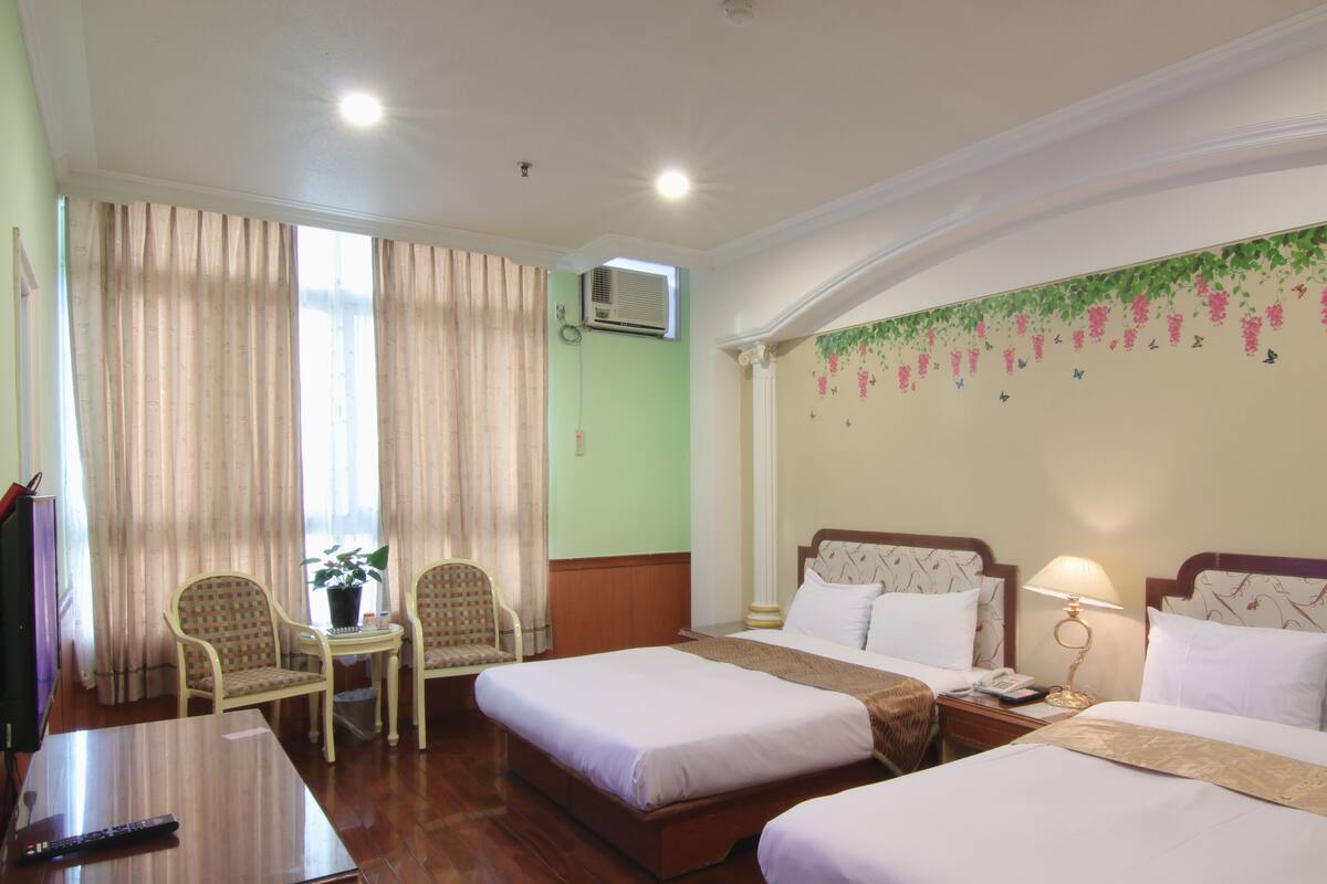 A hotel room with two beds, a desk, a chair, a TV, and a window with curtains.