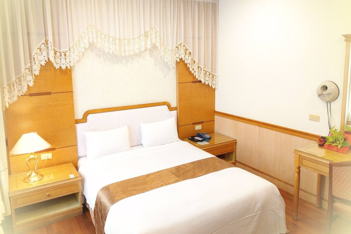 A hotel room with a wooden headboard, a bed with white linens, a nightstand with a lamp, a bedside table with a phone and a plant, and a wall-mounted mirror.