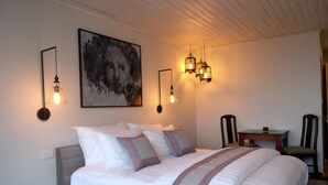 A neatly made bed with a white and gray striped blanket, a painting of a dog on the wall, and a table with chairs in the corner. - The Hill Station Boutique Hotel (Sa Pa)