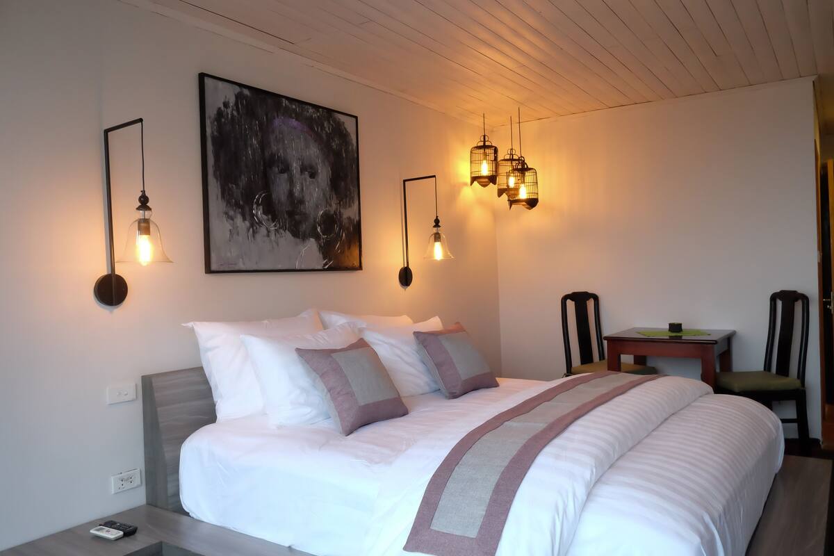 A neatly made bed with a white and gray striped blanket, a painting of a dog on the wall, and a table with chairs in the corner.