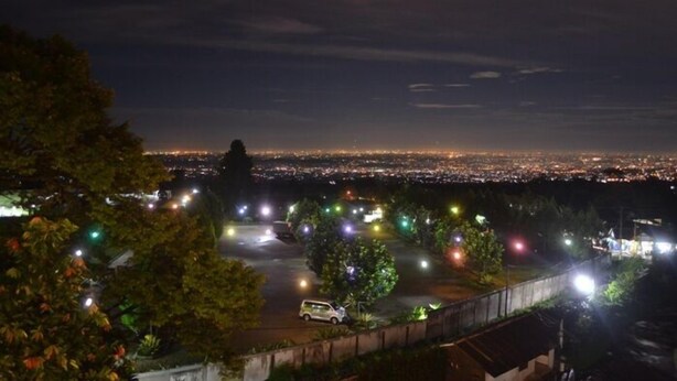 A nighttime view of a cityscape with a distant city skyline, a parking area with trees, and a few buildings in the foreground. - The Highland Park Resort Hotel Bogor (Cilobak)