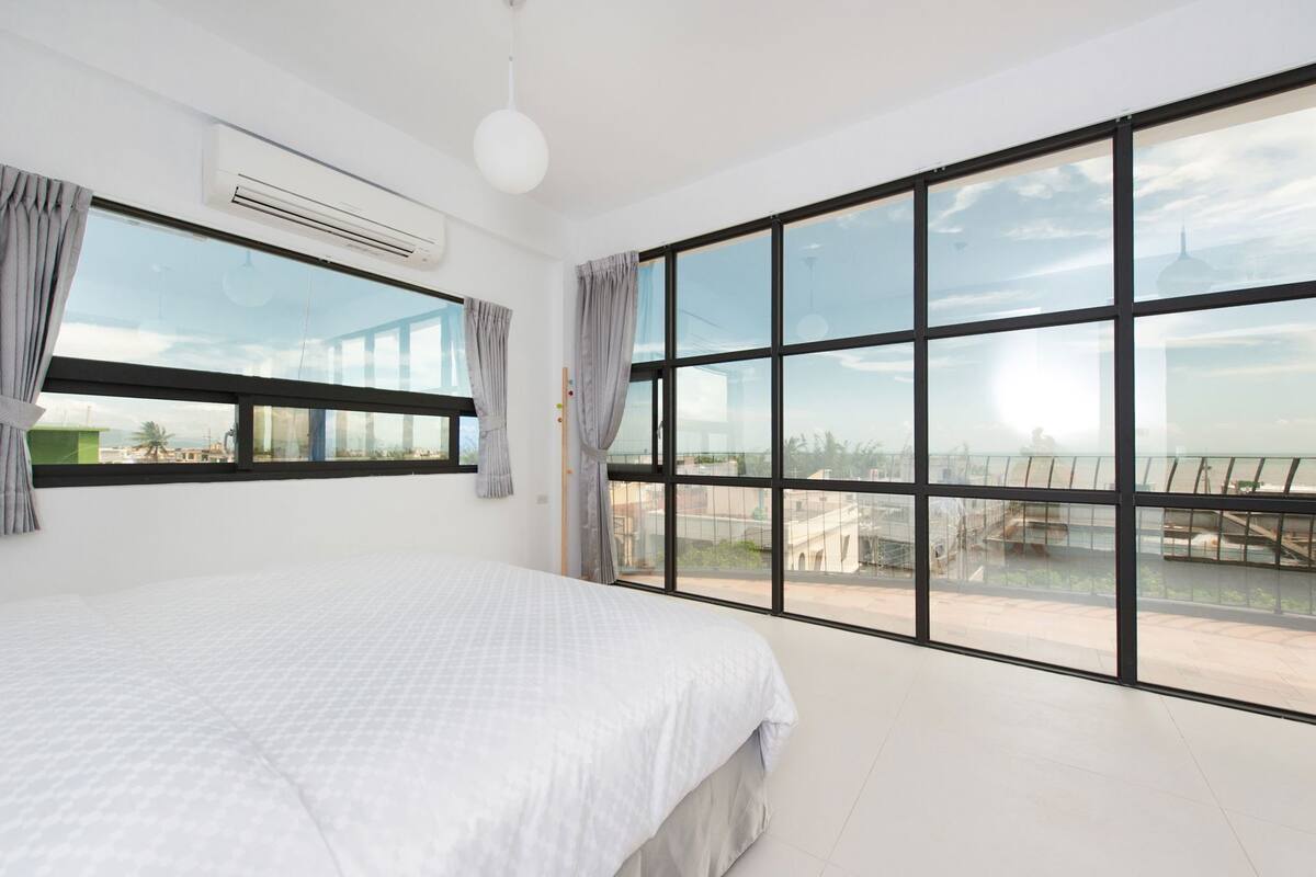A bedroom with a large bed, a window with curtains, an air conditioning unit, and a view of buildings.