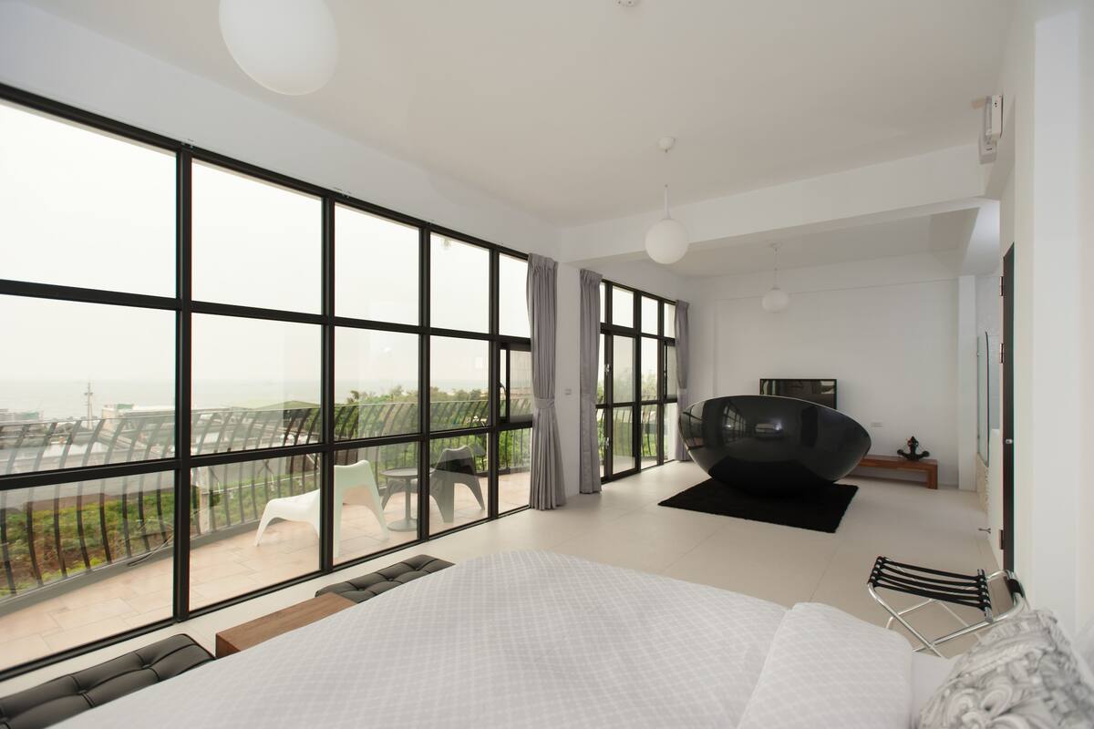 A modern bedroom with a large bed, a view of the city through floor-to-ceiling windows, and a balcony with outdoor seating.