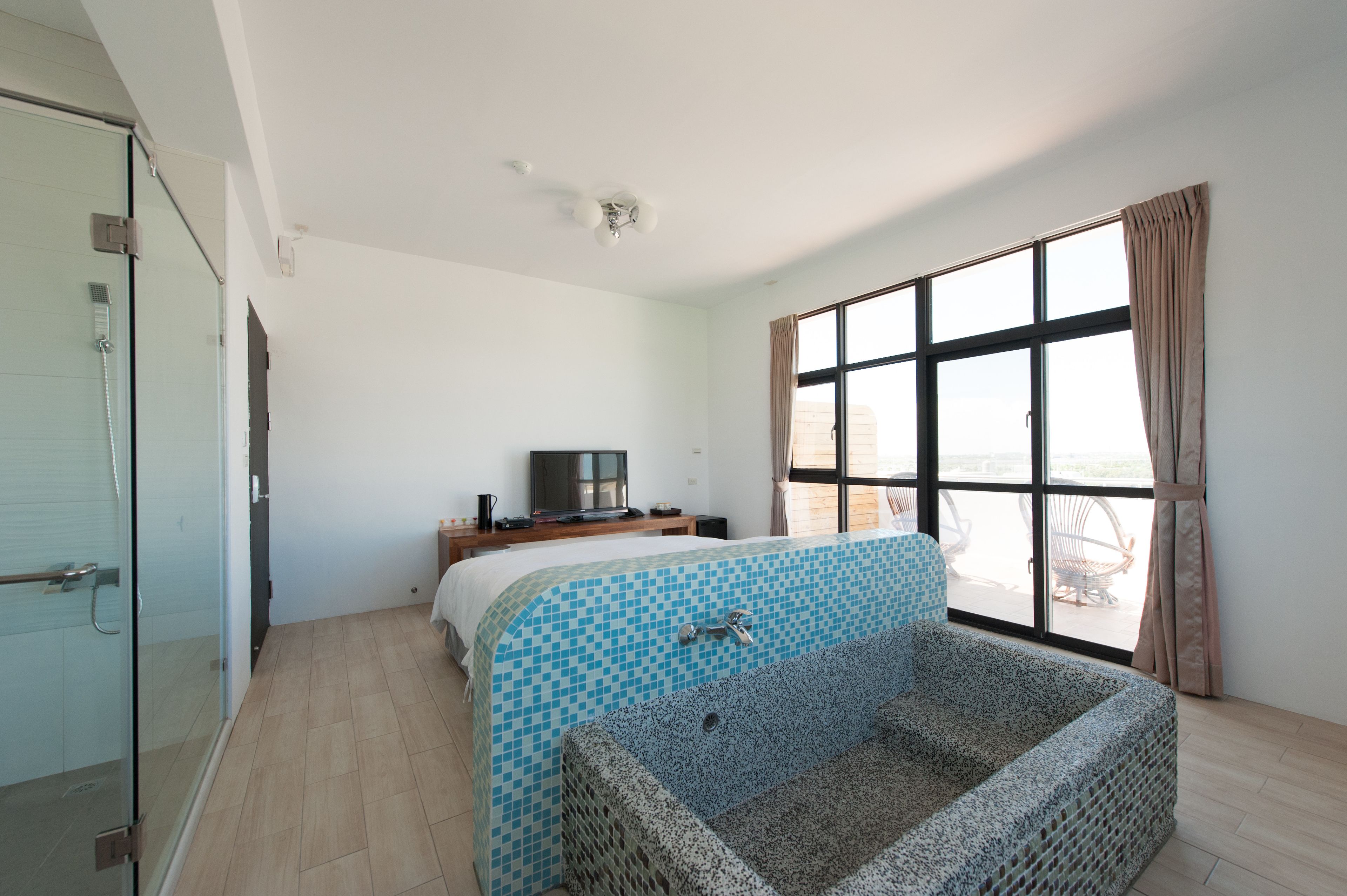 A modern hotel room with a large window, a bathtub, a television, and a glass shower enclosure.