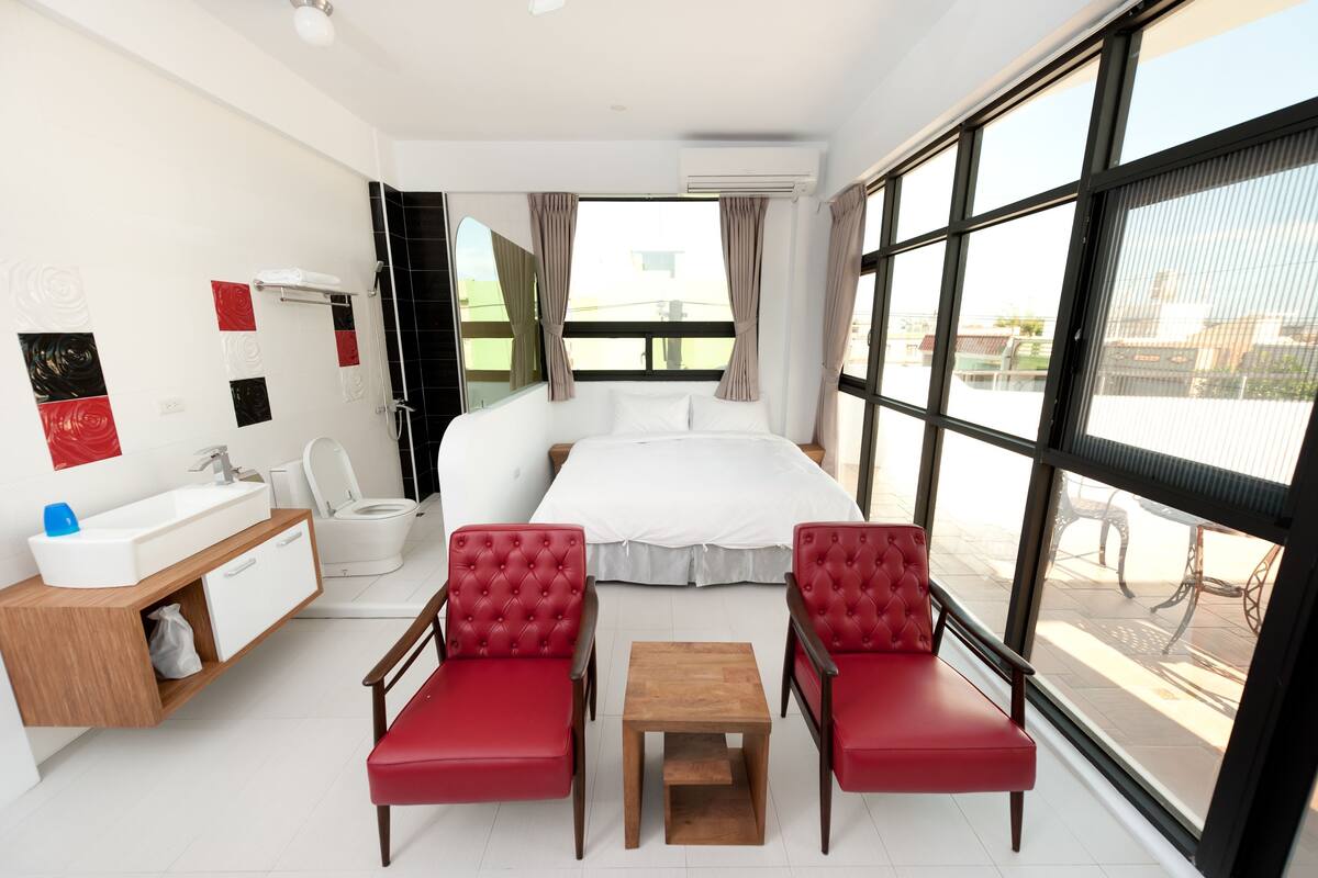 A hotel room with a bed, a bathroom, a small table, two red chairs, and large windows.