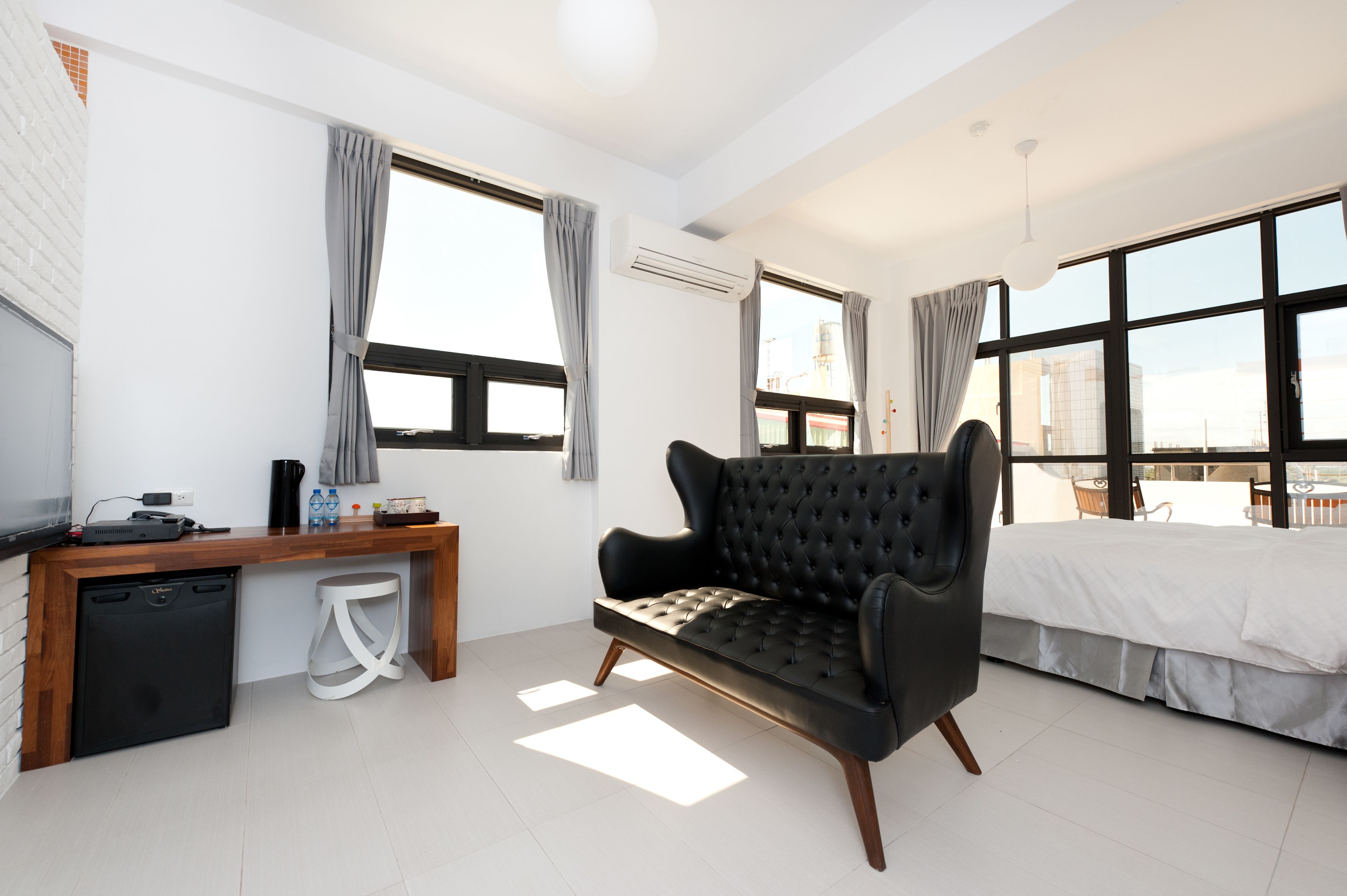 A modern hotel room with a black sofa, a wooden desk, a small fridge, and a bed with white bedding.