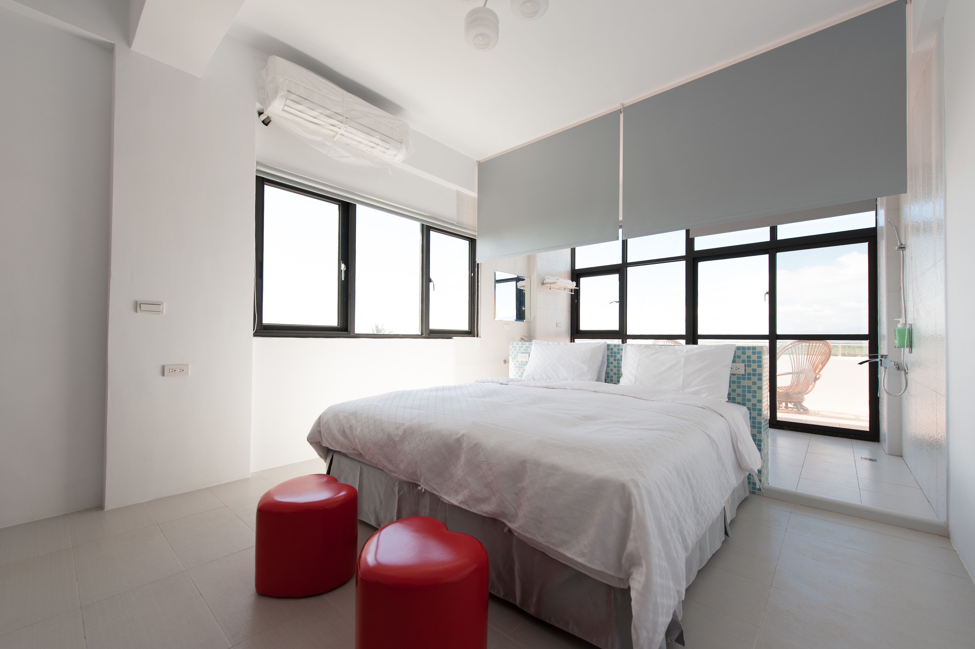 A modern bedroom with a large bed, two red stools, and multiple windows providing natural light.