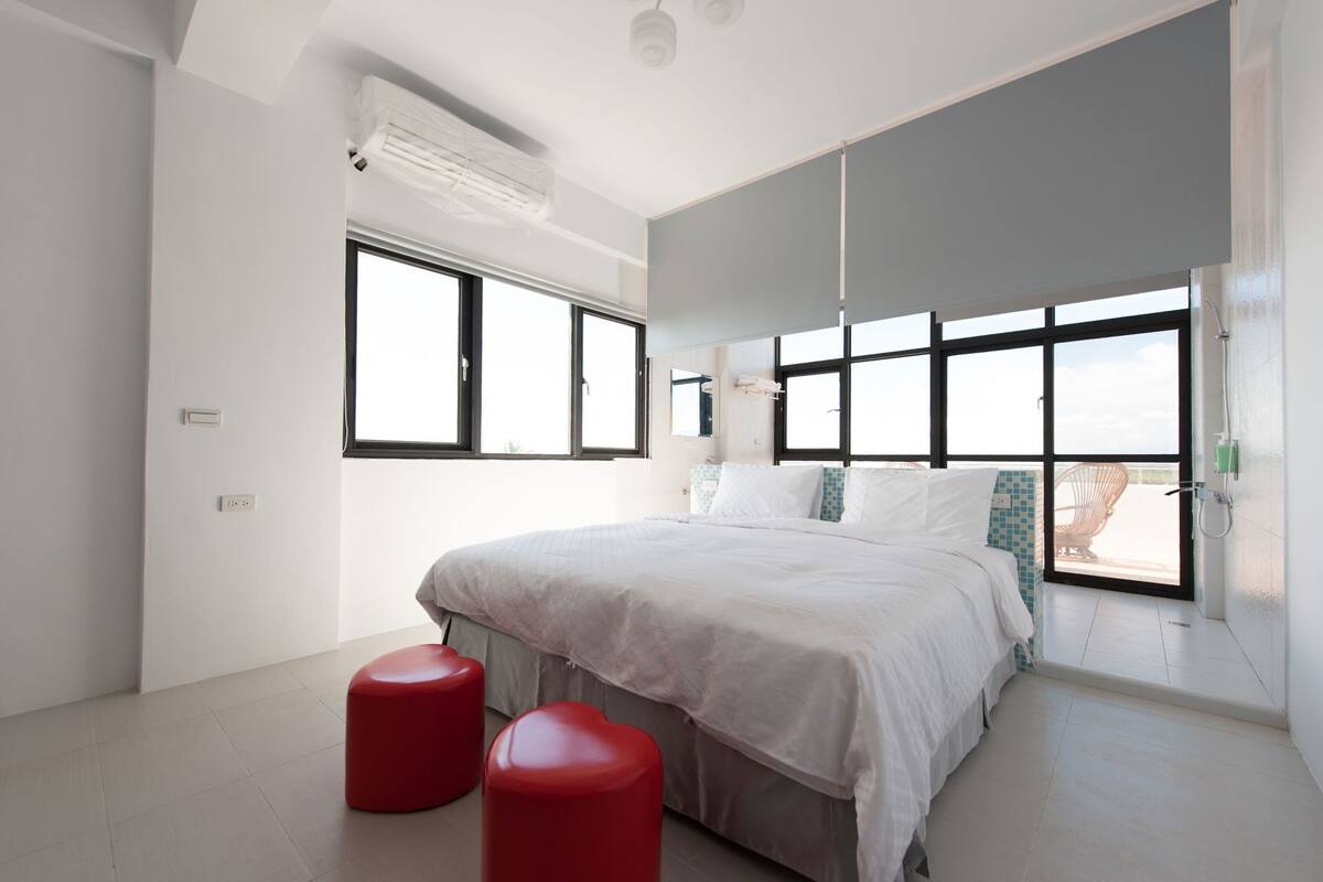 A modern bedroom with a large bed, two red stools, and multiple windows.