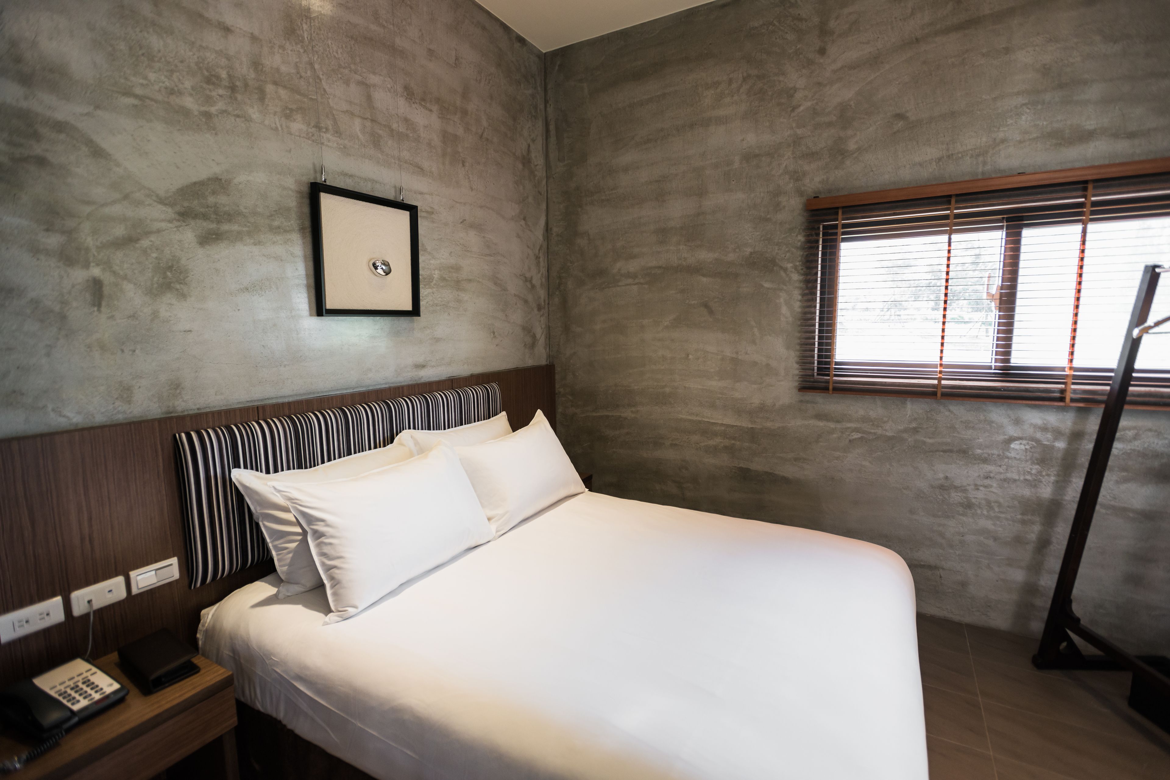 Une chambre d’hôtel avec un grand lit, une table de chevet avec un téléphone, un tableau encadré au mur et une fenêtre avec des stores.