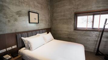 Une chambre d’hôtel avec un grand lit, une table de chevet avec un téléphone, un tableau encadré au mur et une fenêtre avec des stores.