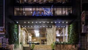 Een moderne hotelentree met een opvallend 'ALANA'-bord, een trap naar de lobby en een goed verlichte gevel.