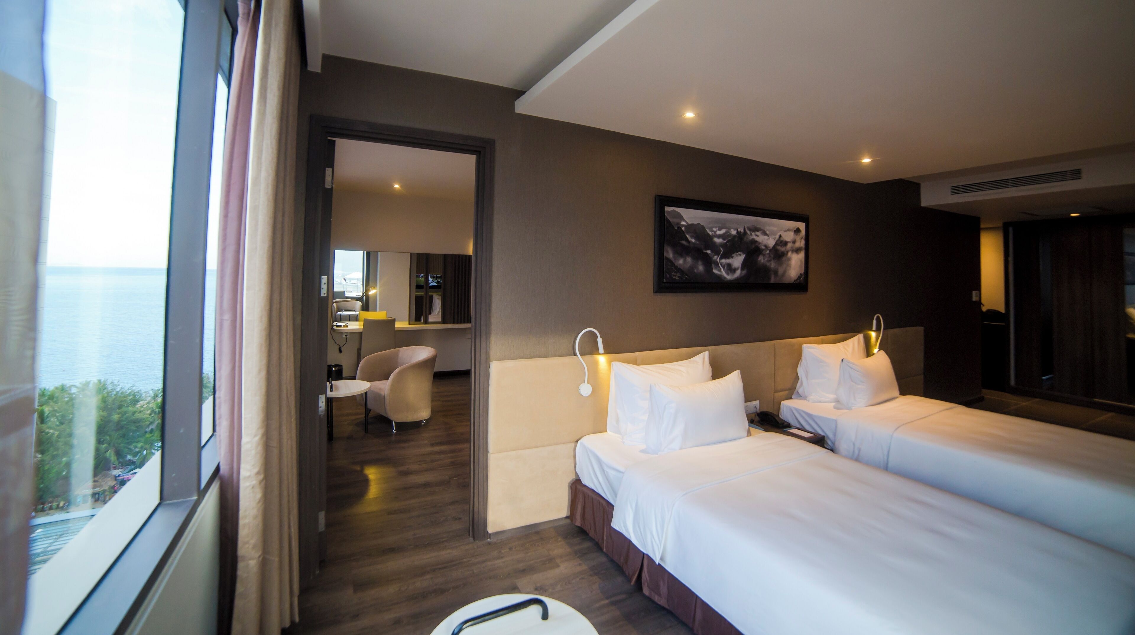 A hotel room with two beds, a desk, and a chair. There is a view of the sea through the window.