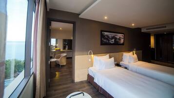 A hotel room with two beds, a desk, and a chair. There is a view of the sea through the window.