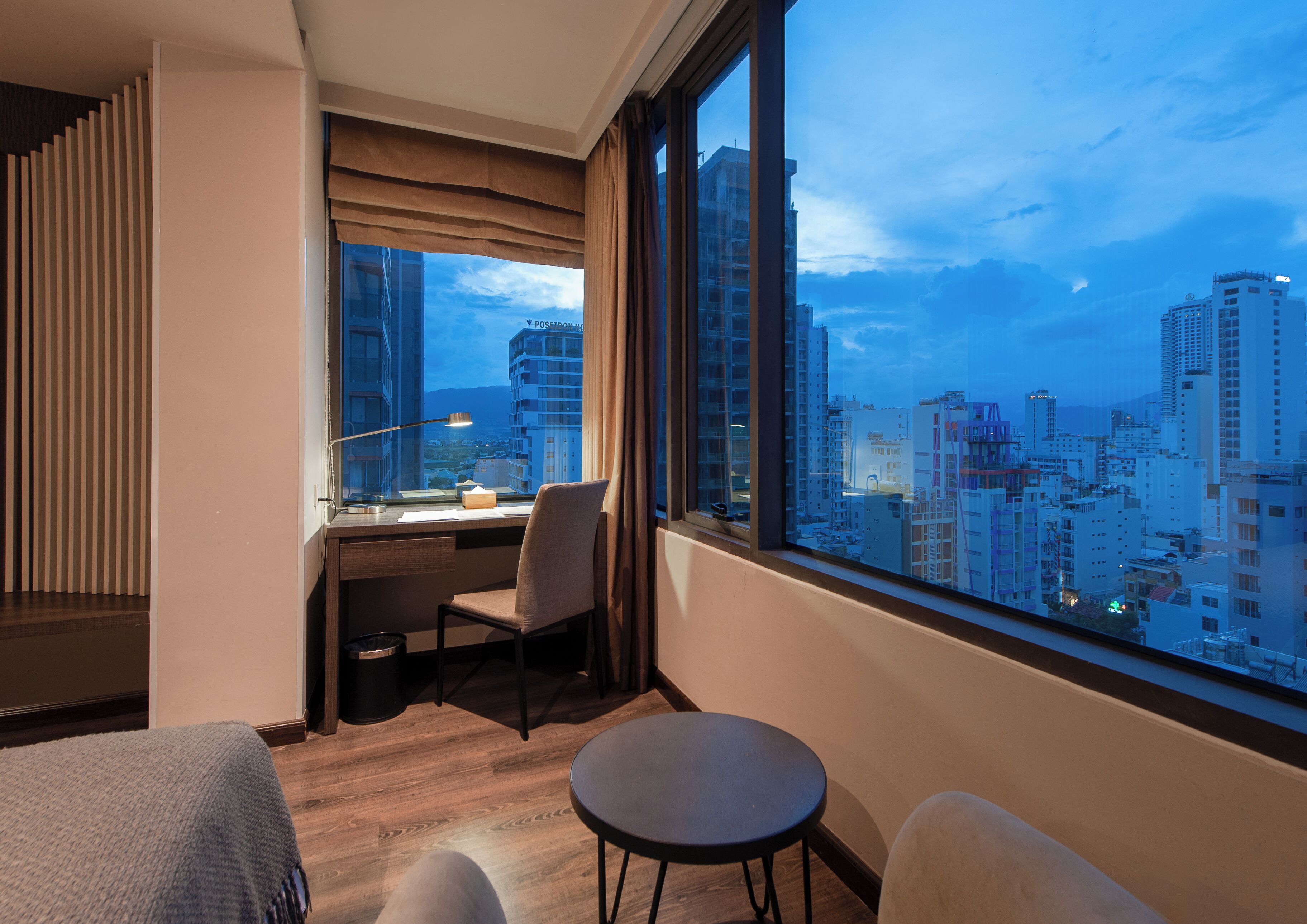 A modern hotel room with a large window offering a city view, a desk with a chair, and a small round table.