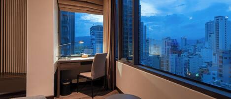 A modern hotel room with a large window offering a city view, a desk with a chair, and a small round table.