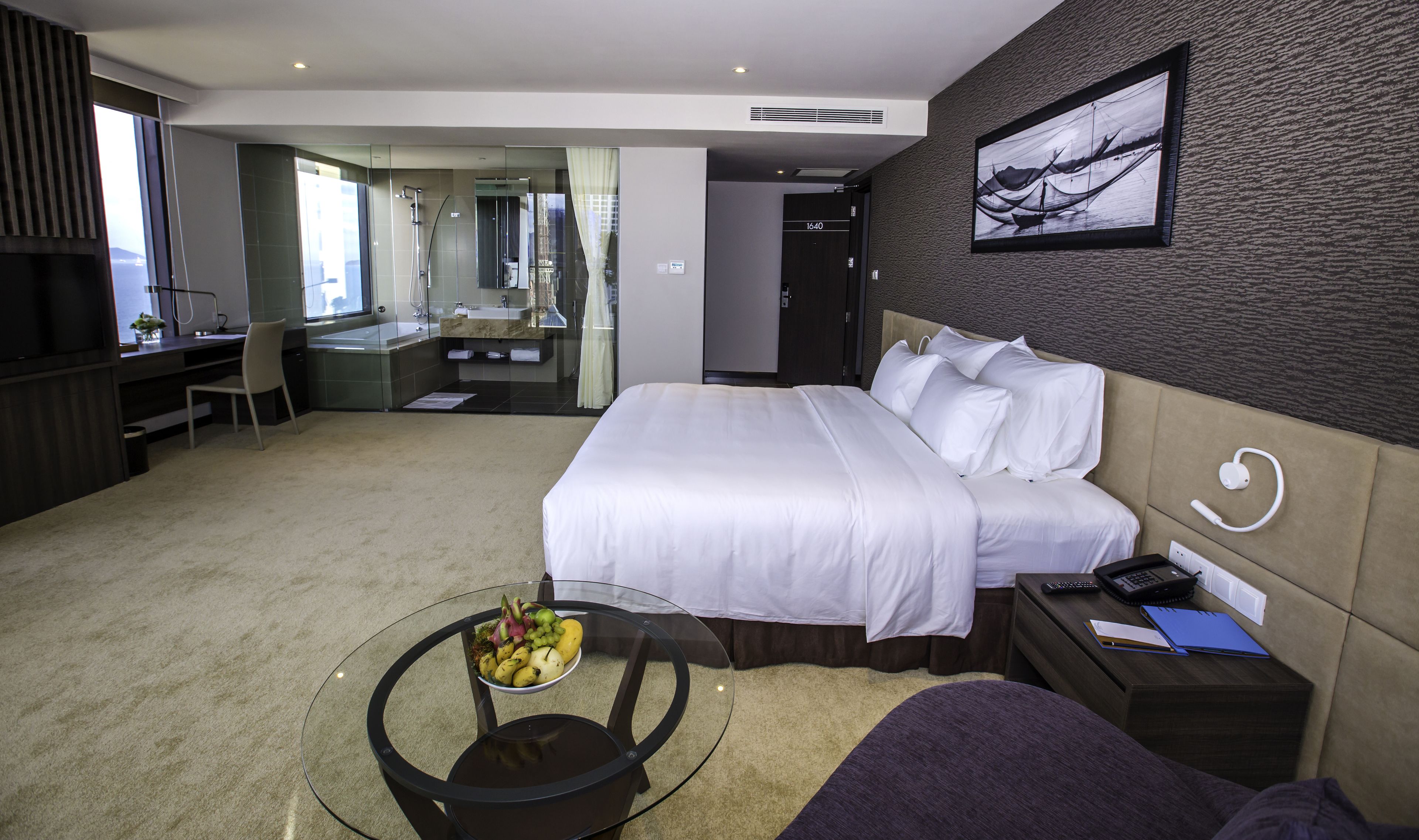 Une chambre d’hôtel moderne avec un grand lit, un bureau et une table en verre sur laquelle sont disposés des fruits.