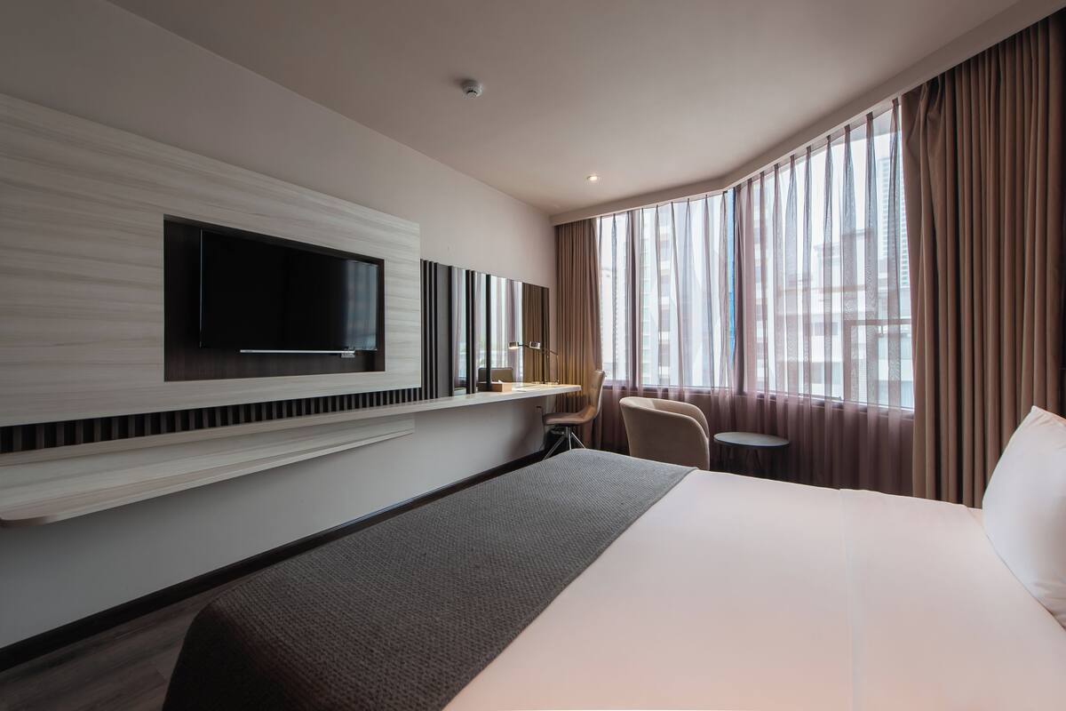 Habitación de hotel con una cama grande, un televisor de pantalla plana montado en la pared, un escritorio con una silla y un ventanal con cortinas.