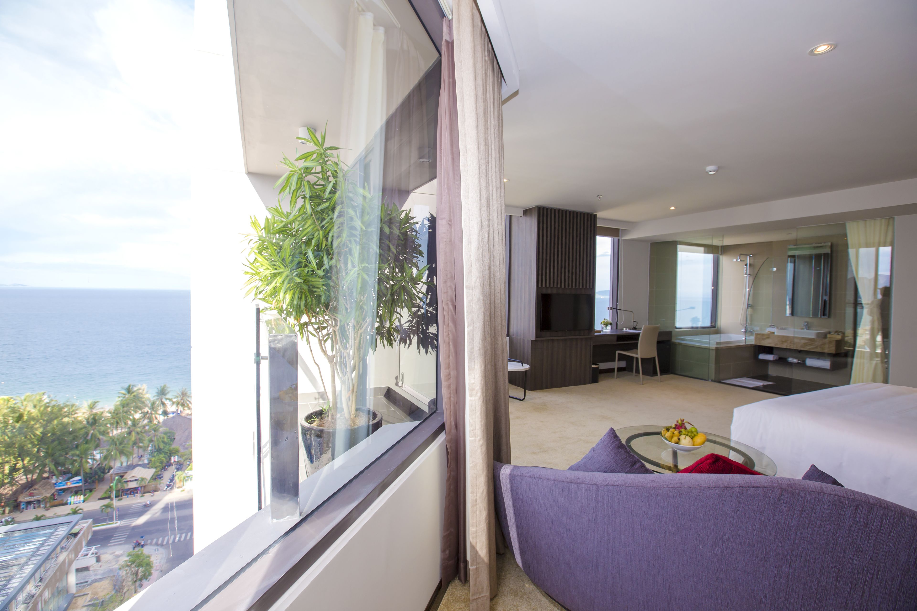 A modern hotel room with a large window overlooking the ocean, a bed, a seating area, and a kitchenette.
