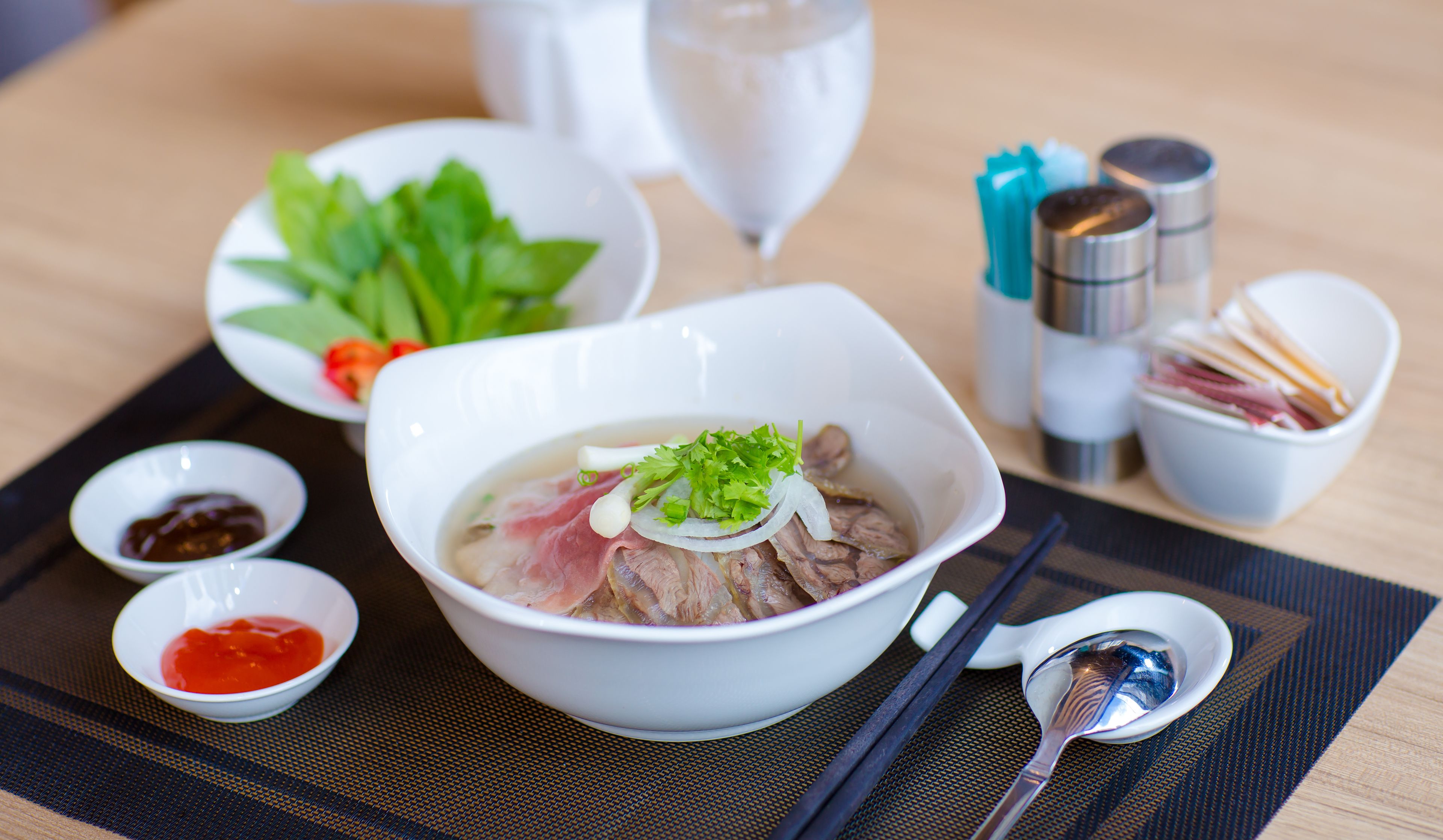 A bowl of soup with meat and vegetables, accompanied by small bowls of condiments and chopsticks on a table.