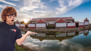 A person standing in front of a building with a red roof and white walls, reflected in a calm body of water.
